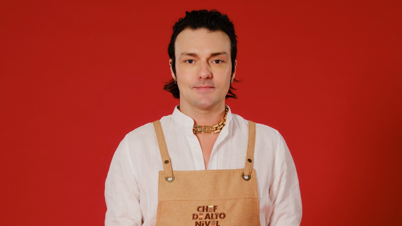 Lucio Flajore posa com camisa branca, colar dourado e avental bege de Chef de Alto Nível, diante de um fundo vermelho
