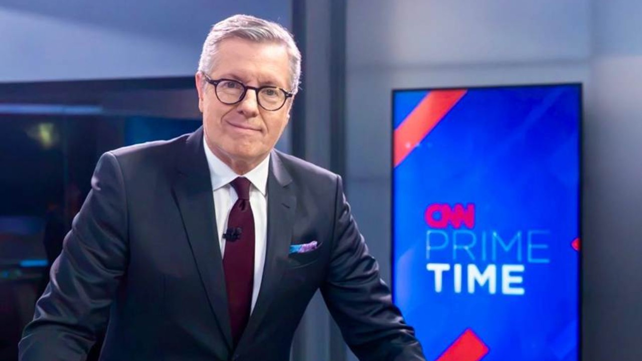 Márcio Gomes aparece de terno escuro, gravata vinho e óculos no estúdio da CNN Brasil, ao lado de uma tela com o logotipo do CNN Prime Time