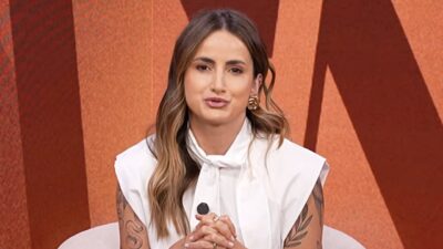Mari Palma aparece em estúdio de televisão, com blusa branca, cabelos soltos, brincos dourados e tatuagens nos braços, diante de um cenário em tons alaranjados