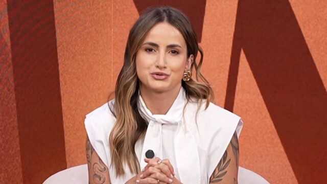 Mari Palma aparece em estúdio de televisão, com blusa branca, cabelos soltos, brincos dourados e tatuagens nos braços, diante de um cenário em tons alaranjados