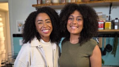 Mariana Bispo e Thallita Flor sorriem lado a lado em uma cozinha, com utensílios e potes ao fundo