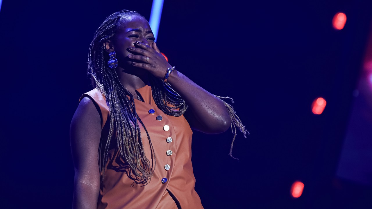 Mariana Lourenço leva uma mão à boca no palco do The Voice Brasil, com tranças longas e roupa marrom com botões metálicos