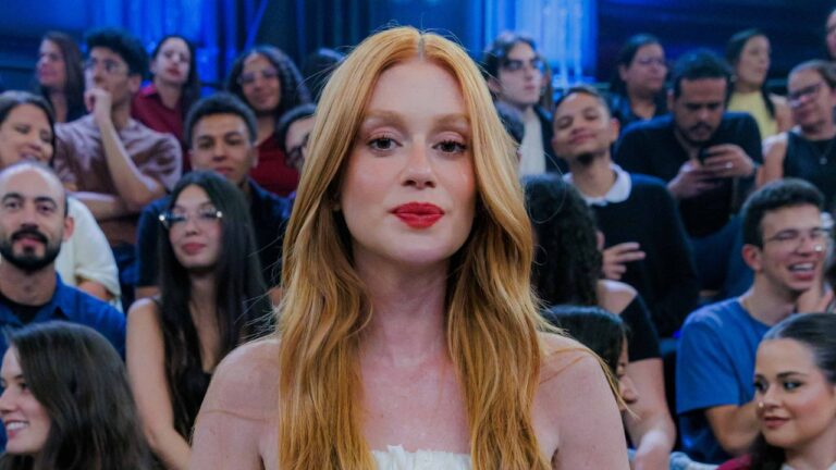 Marina Ruy Barbosa aparece em primeiro plano, com cabelos ruivos longos e soltos, batom vermelho e roupa branca, diante de uma plateia em um estúdio de televisão com iluminação azul