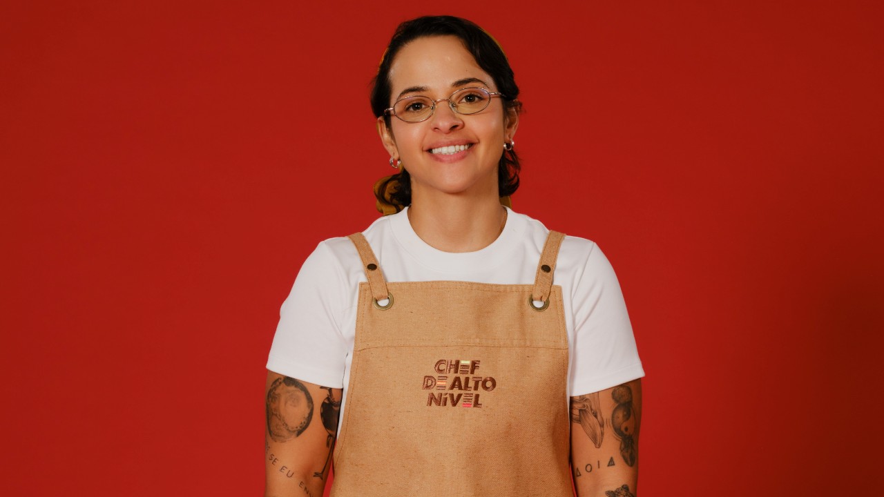 Marina Siqueira sorri com óculos, camiseta branca e avental bege de Chef de Alto Nível, com tatuagens nos braços e fundo vermelho