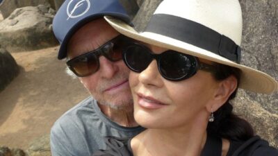 Michael Douglas e Catherine Zeta-Jones posam juntos ao ar livre, com óculos escuros; ele usa boné azul e ela, chapéu claro com faixa preta