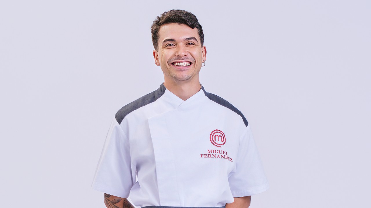 Miguel Fernandez sorri com uniforme branco do MasterChef Profissionais diante de fundo claro