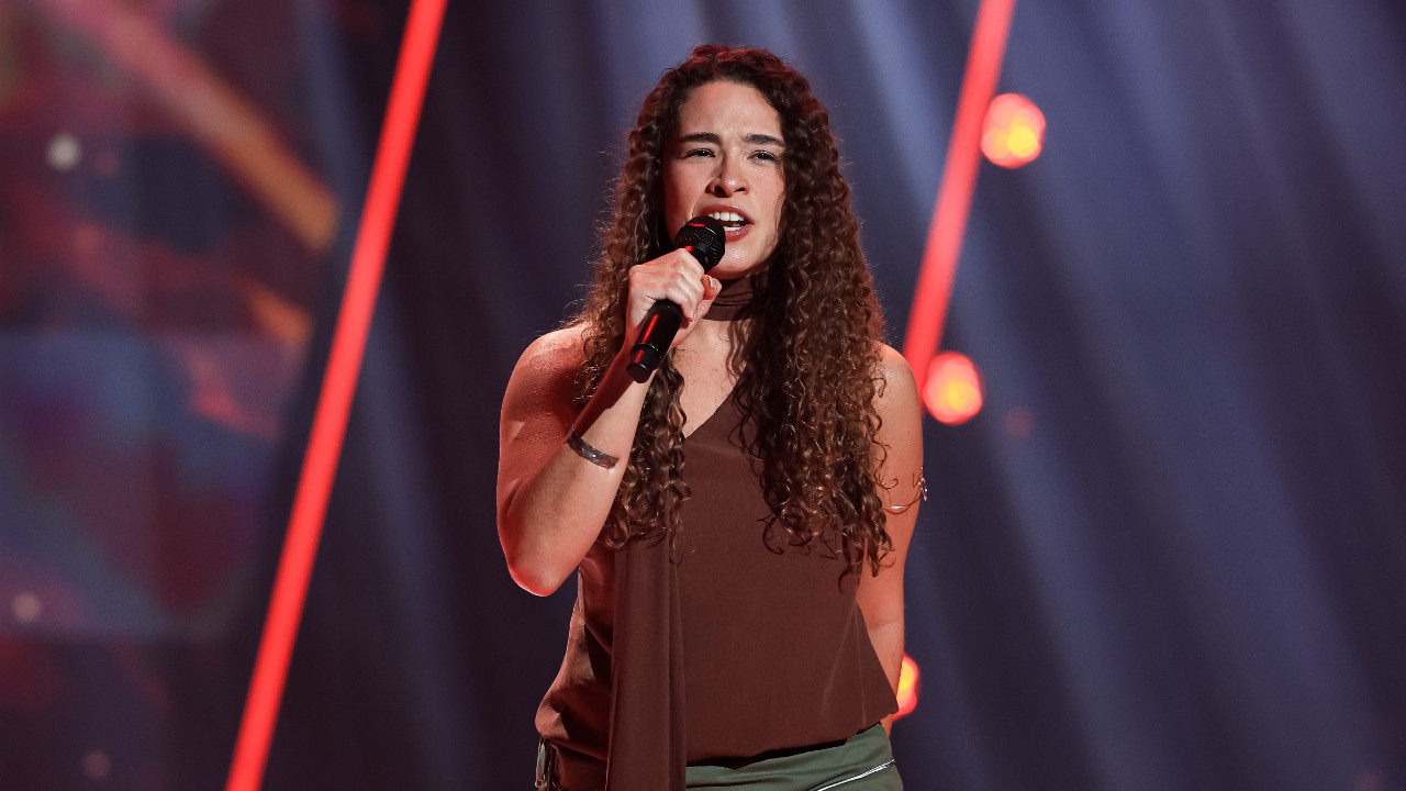 Milena Amaral canta ao microfone com blusa marrom e cabelos cacheados, diante de luzes vermelhas no palco