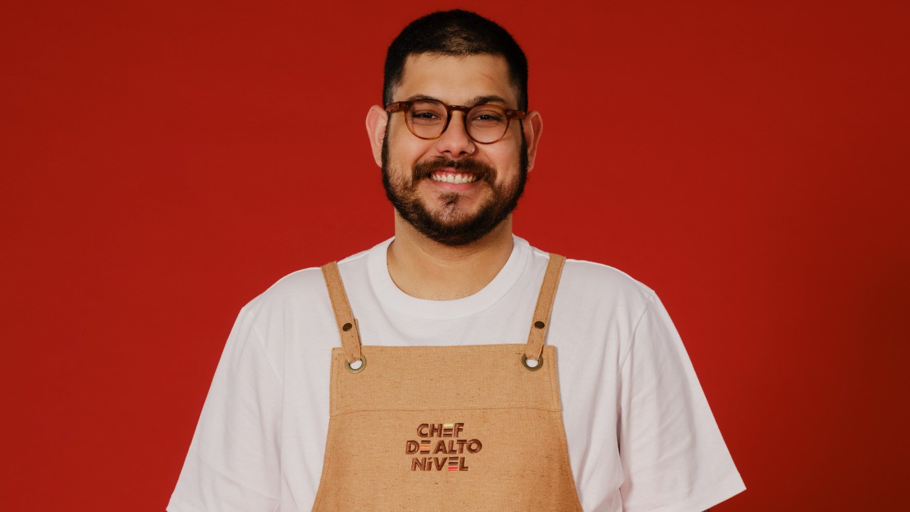 Mitchell Figueiredo sorri com óculos marrons, barba, camiseta branca e avental bege de Chef de Alto Nível, diante de um fundo vermelho