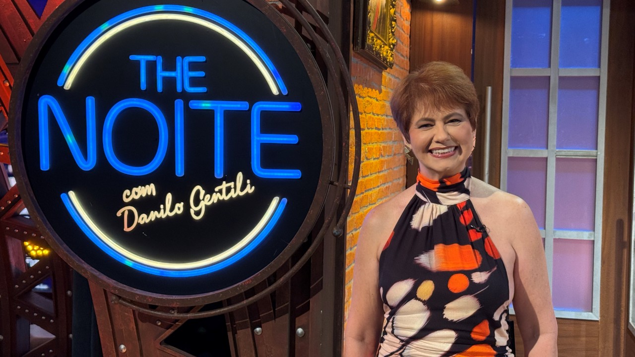Narjara Turetta aparece sorridente ao lado do letreiro iluminado do The Noite, no SBT
