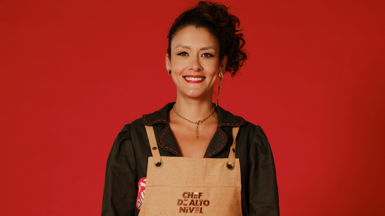 Nathascha Francis sorri com cabelo preso de lado, blusa escura e avental bege de Chef de Alto Nível, diante de um fundo vermelho