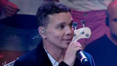 Netinho aparece com cabelos curtos, veste casaco escuro e segura um microfone durante apresentação musical, com integrantes da banda ao fundo