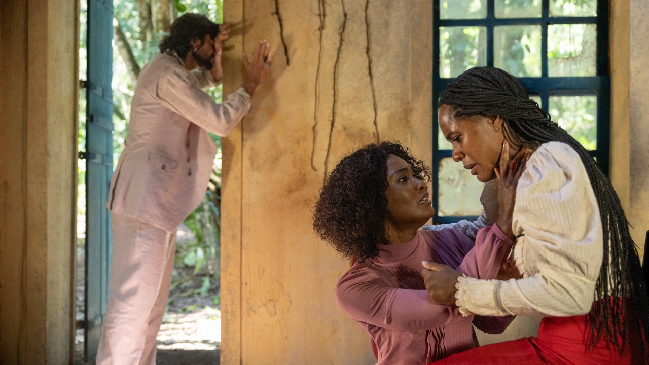 Omar (Rodrigo Simas) aparece ao fundo, enquanto Niara (Erika Januza), de roupa rosa, ampara Alika (Duda Santos), que veste blusa branca e saia vermelha, em uma casa rústica
