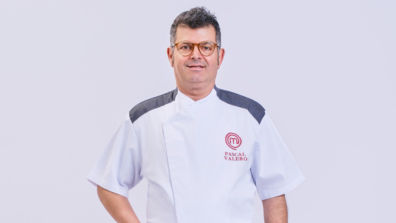 Pascal Valero posa de óculos e uniforme branco do MasterChef Profissionais diante de fundo claro