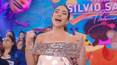 Patricia Abravanel sorri com as mãos erguidas, em vestido rosa com brilho, diante da plateia e do cenário colorido do Programa Silvio Santos