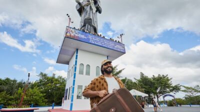 Paulo Vieira sorri com uma mala marrom nas mãos diante de uma torre com estátua religiosa, em uma praça ao ar livre