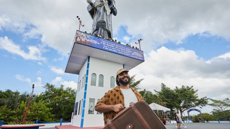 Paulo Vieira sorri com uma mala marrom nas mãos diante de uma torre com estátua religiosa, em uma praça ao ar livre