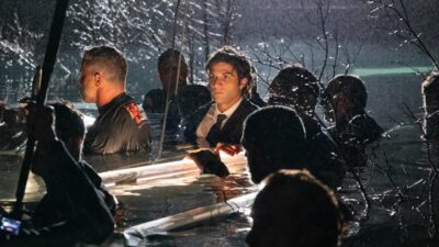 Pedro (Chay Suede) aparece de terno e gravata, com os cabelos molhados, dentro de um rio durante uma cena noturna, cercado por pessoas na água e sob iluminação de equipamentos de gravação