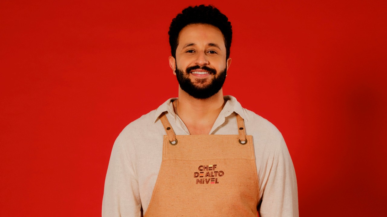 Pedro Fartura sorri com barba, camisa clara e avental bege de Chef de Alto Nível, diante de um fundo vermelho
