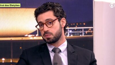 Pedro Figueiredo em close no estúdio da Central das Eleições, com óculos de armação marrom, terno escuro e gravata cinza