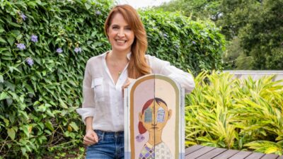 Poliana Abritta sorri em um jardim, com camisa branca e calça jeans, ao lado de um painel ilustrado com um rosto