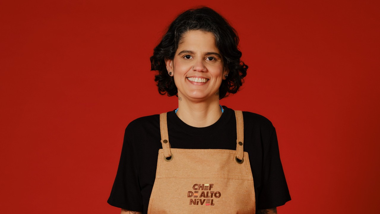 Rafaela Cabadas sorri com camiseta preta e avental bege de Chef de Alto Nível, diante de um fundo vermelho