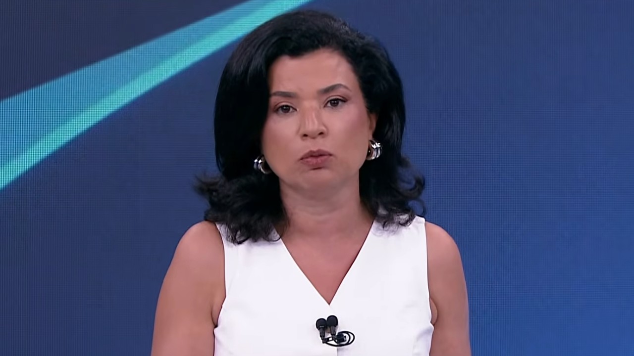 Raquel Landim de blusa branca sem mangas e brincos prateados diante de painel azul em estúdio de televisão