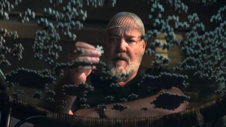 Russell Crowe como Roy Freeman em A Teia, com uma peça de quebra-cabeça na mão diante de peças mesa de vidro