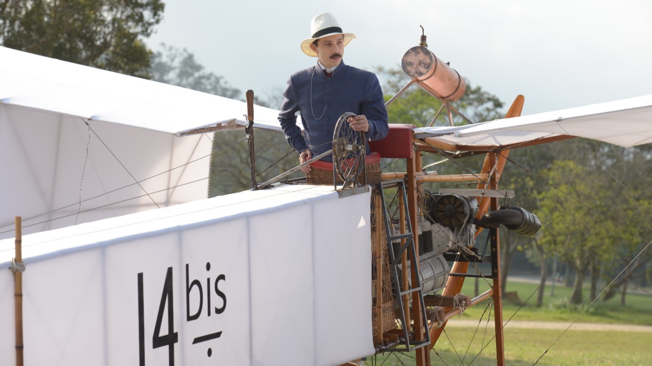 João Pedro Zappa interpreta Santos Dumont em cena da minissérie, usando chapéu branco e traje azul enquanto aparece na estrutura de uma réplica do avião 14-bis, com asas de tecido, cabos e motor expostos