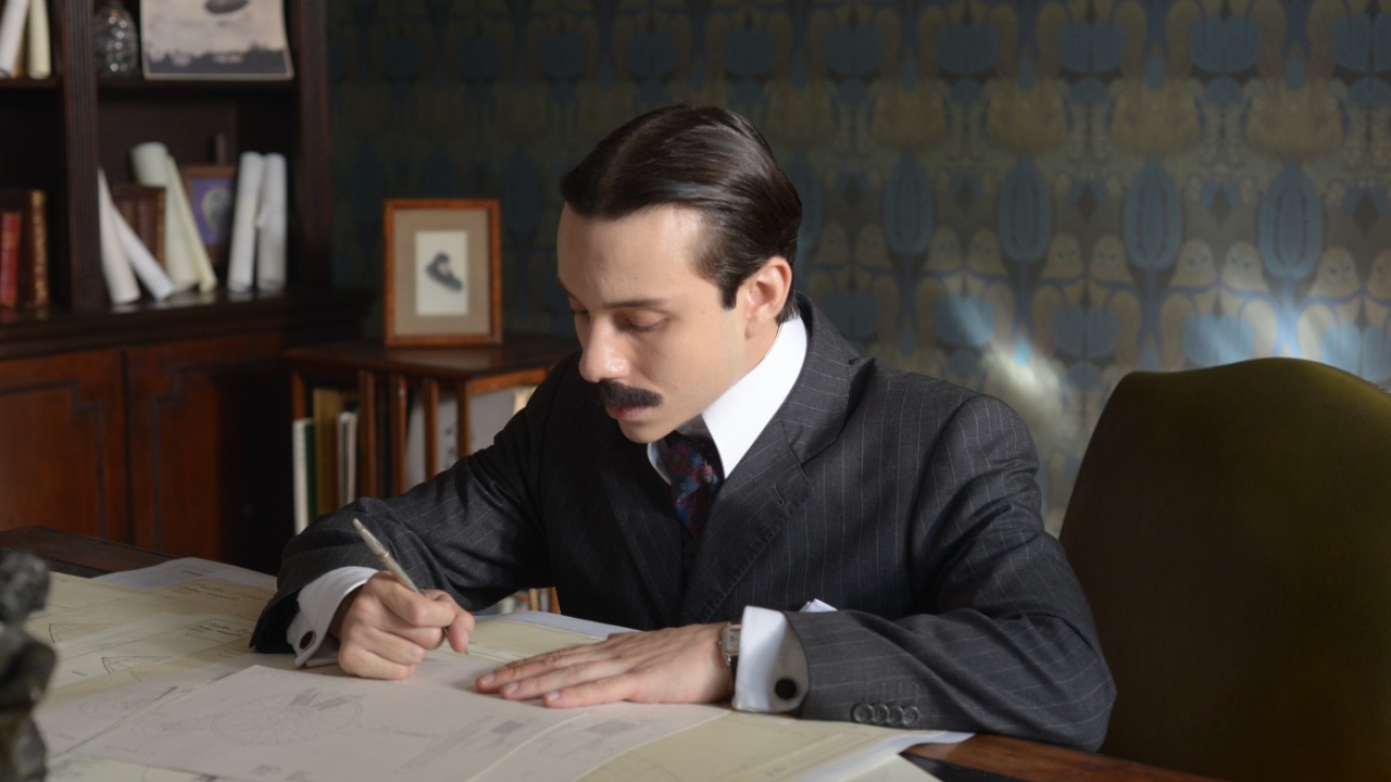 João Pedro Zappa interpreta Santos Dumont em cena da minissérie, sentado à mesa, com terno escuro e bigode, enquanto escreve sobre desenhos técnicos em um ambiente de época