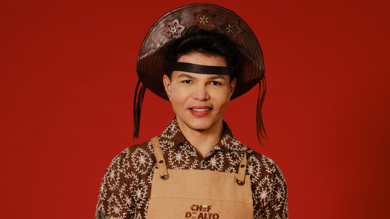 Stenio Birth posa com chapéu de cangaceiro, camisa marrom estampada e avental bege de Chef de Alto Nível, diante de um fundo vermelho