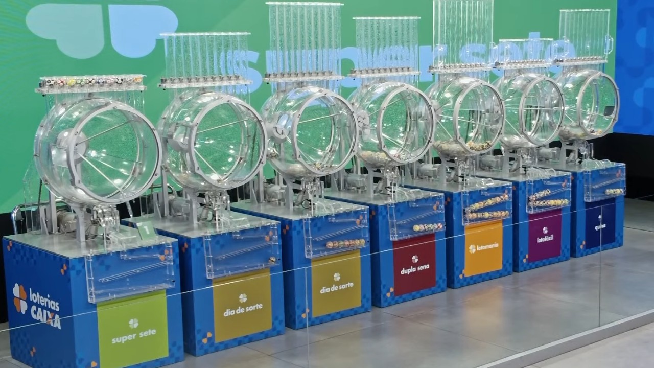 Sete globos transparentes de sorteio alinhados sobre bases azuis das Loterias Caixa, com bolinhas numeradas em compartimentos e um painel verde ao fundo com a identificação do Super Sete