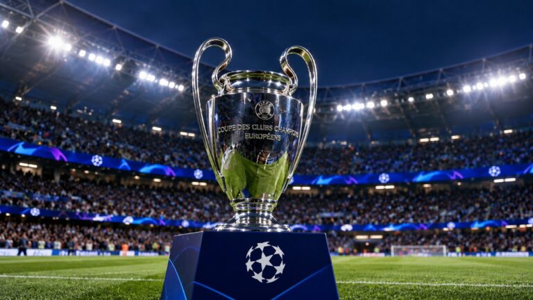 Taça da UEFA Champions League exposta sobre um pedestal no gramado de um estádio de futebol, com arquibancadas ocupadas por torcedores ao fundo