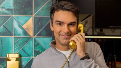 Tadeu Schmidt sorri com um telefone dourado junto ao ouvido, diante de uma parede com revestimento geométrico azul e dourado