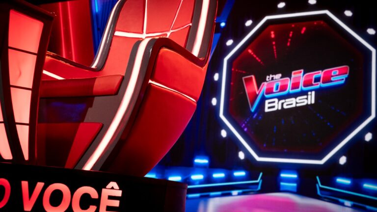 Cadeira vermelha do The Voice Brasil em primeiro plano com logotipo do programa exibido no telão do palco