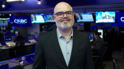 Thiago Contreira de terno escuro e óculos sorri diante de câmeras e monitores na redação da CNBC