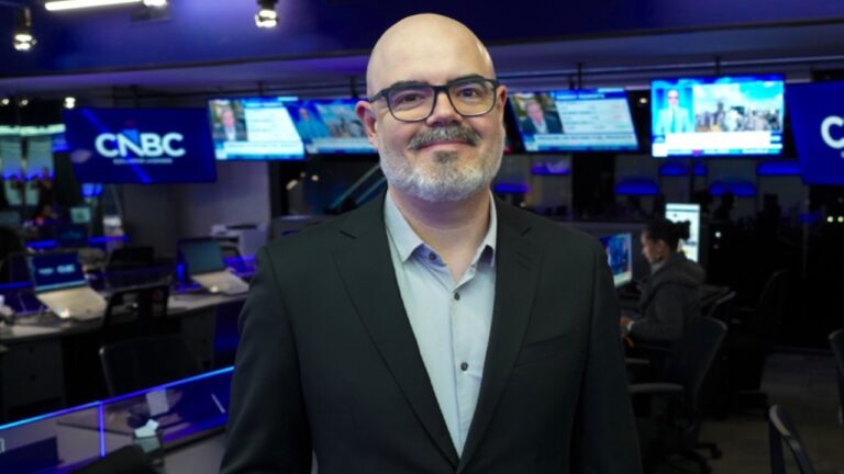 Thiago Contreira de terno escuro e óculos sorri diante de câmeras e monitores na redação da CNBC