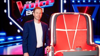 Tiago Leifert posa com terno azul e camiseta branca ao lado de uma cadeira vermelha, com o logotipo do The Voice Brasil ao fundo