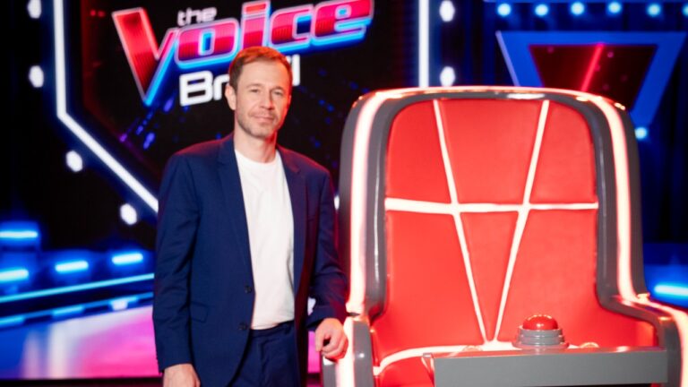 Tiago Leifert posa com terno azul e camiseta branca ao lado de uma cadeira vermelha, com o logotipo do The Voice Brasil ao fundo