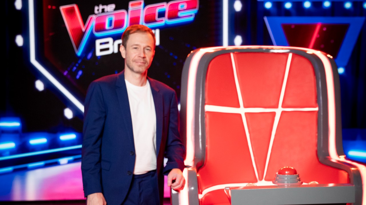 Tiago Leifert posa com terno azul e camiseta branca ao lado de uma cadeira vermelha, com o logotipo do The Voice Brasil ao fundo