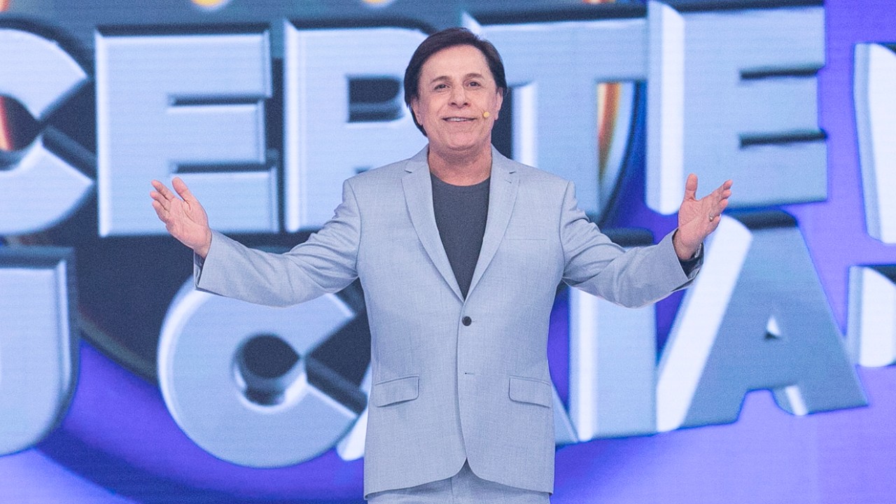 Tom Cavalcante aparece de braços abertos no palco do Acerte ou Caia!, da Record, diante do cenário do programa, vestindo terno azul-claro
