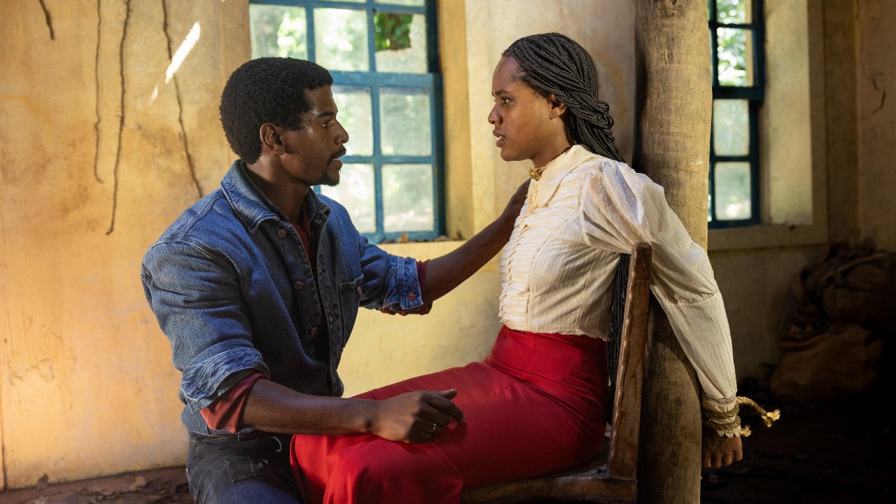 Tonho (Ronald Sotto), de camisa jeans azul, conversa com Alika (Duda Santos), que aparece sentada em uma cadeira, com blusa branca e saia vermelha, em um ambiente rústico