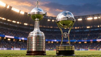 Troféus da Libertadores e da Copa Sul-Americana aparecem lado a lado no gramado de um estádio de futebol iluminado, com arquibancadas ao fundo