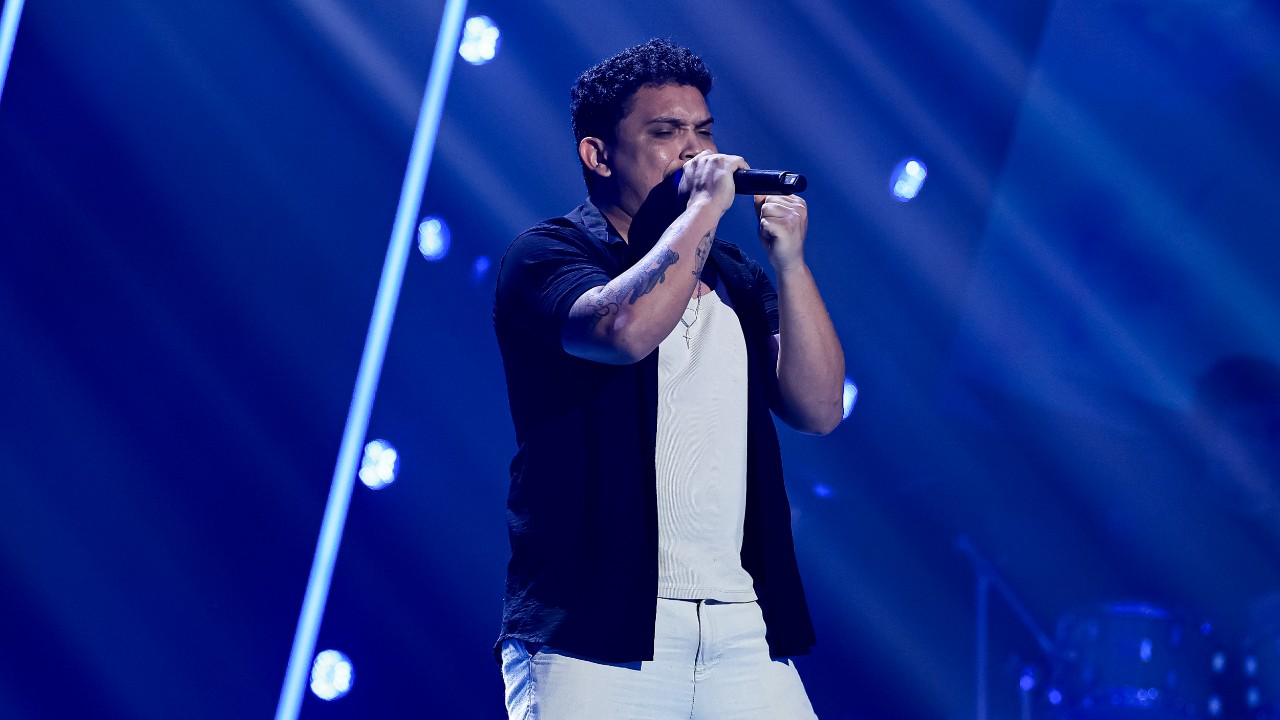 Vini Costta canta ao microfone com camisa preta aberta sobre camiseta branca, uma mão fechada junto ao rosto e iluminação azul