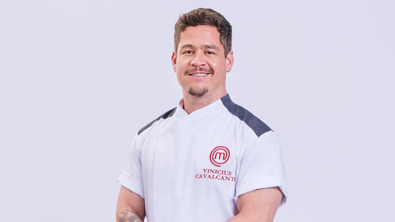 Vinicius Cavalcante sorri com uniforme branco do MasterChef Profissionais diante de fundo claro