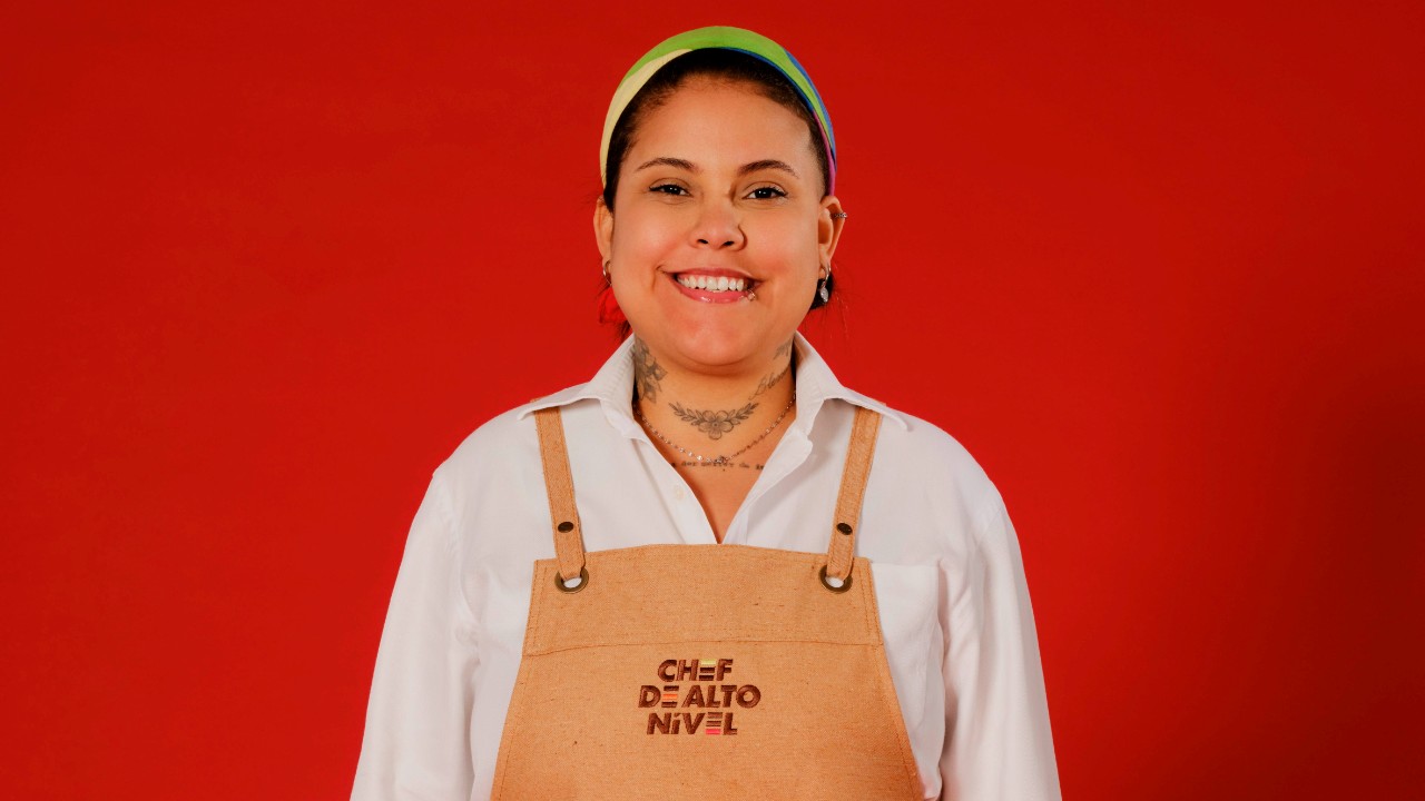 Vitória Cruz sorri com faixa colorida na cabeça, camisa branca e avental bege de Chef de Alto Nível, diante de um fundo vermelho