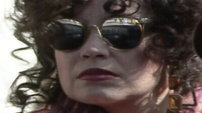 Viúva Porcina (Regina Duarte) de óculos escuros, cabelos cacheados, batom vermelho e brincos dourados em close no rosto
