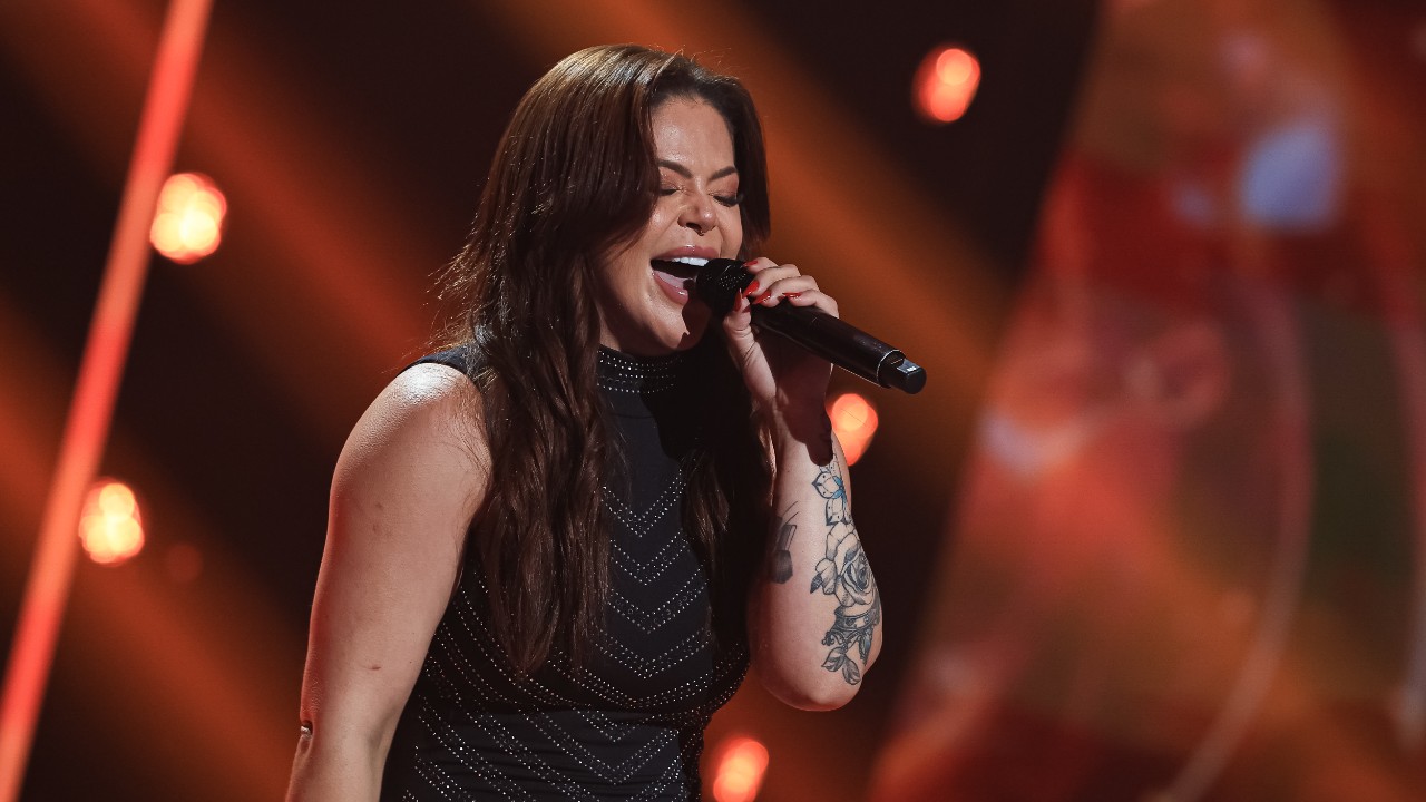 Vivi Alves canta de olhos fechados ao microfone, com roupa preta e tatuagens no braço, sob iluminação vermelha