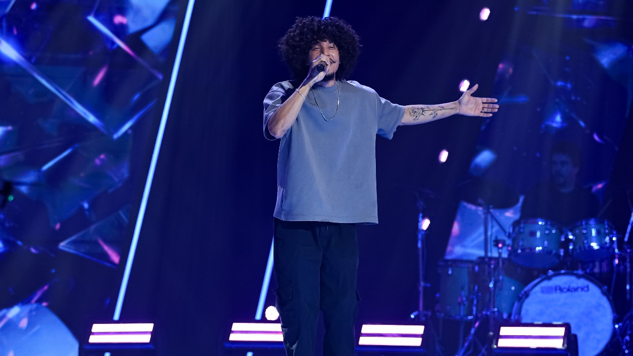Will canta ao microfone com camiseta cinza e braço tatuado estendido, com bateria e luzes azuis ao fundo