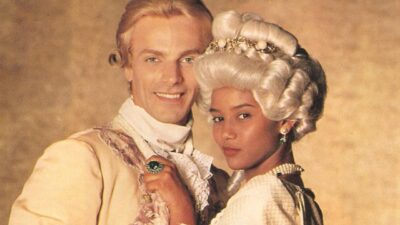 Taís Araujo como Xica e Victor Wagner como João Fernandes aparecem juntos, com roupas de época, perucas e acessórios característicos da novela Xica da Silva