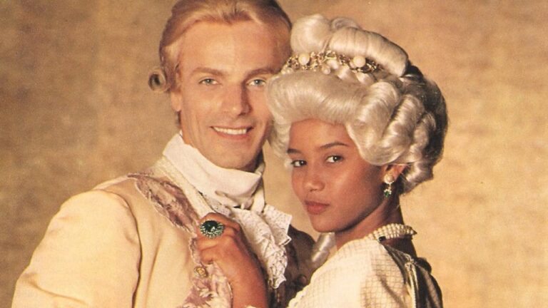 Taís Araujo como Xica e Victor Wagner como João Fernandes aparecem juntos, com roupas de época, perucas e acessórios característicos da novela Xica da Silva